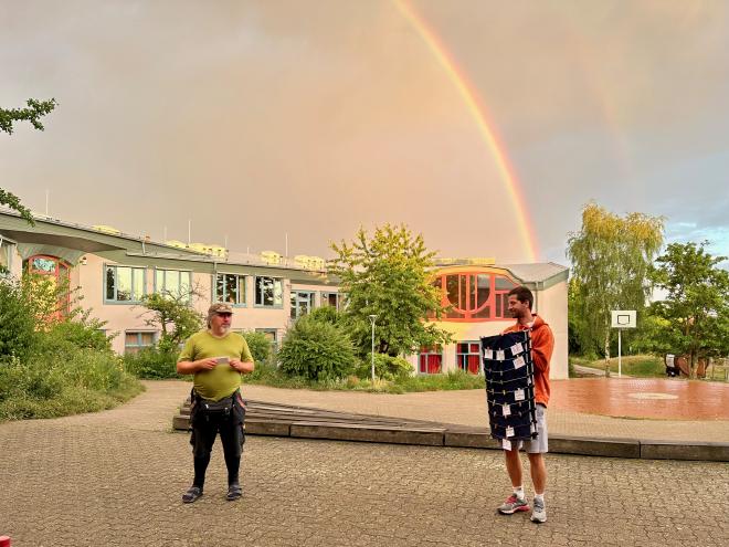 Two men standing in front of a school with a rainbow in the background