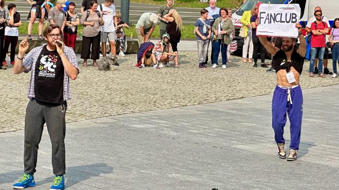 &ldquo;a man holding a microphone and a very fit man holding a “fan club” sign&rdquo;