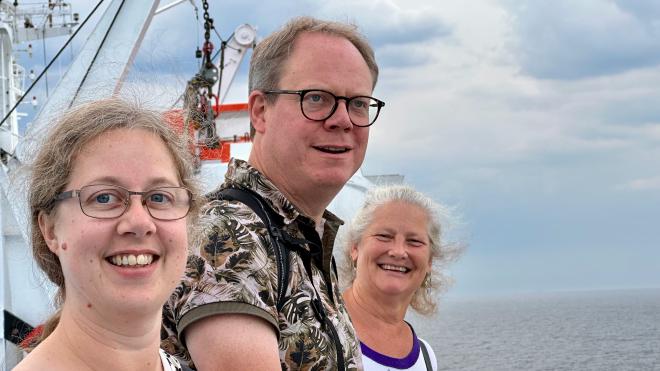 &ldquo;three people standing on a ferry deck with the ocean and sky behind them&rdquo;