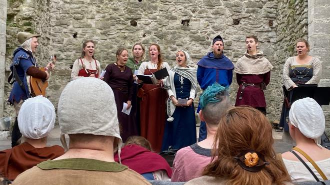 &ldquo;a guitarist and several singers in medieval dress on a stage in front of a stone wall&rdquo;
